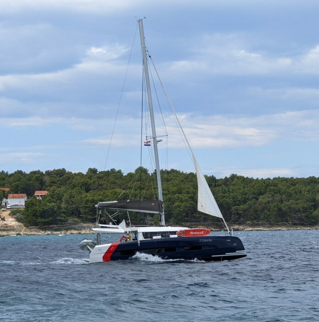 blue catamaran is heading out to the sea while hoisting the genoa
